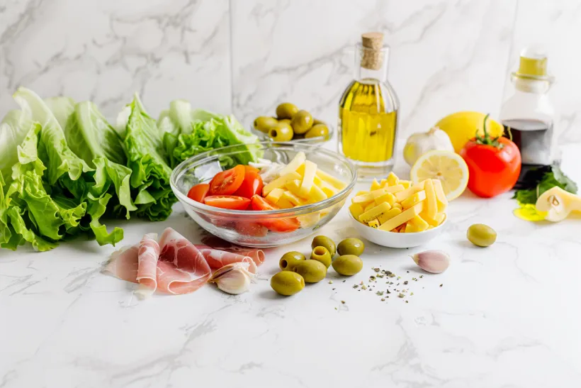 Minimalist side-view of 1905 salad ingredients arranged around a clear glass bowl on white marble.